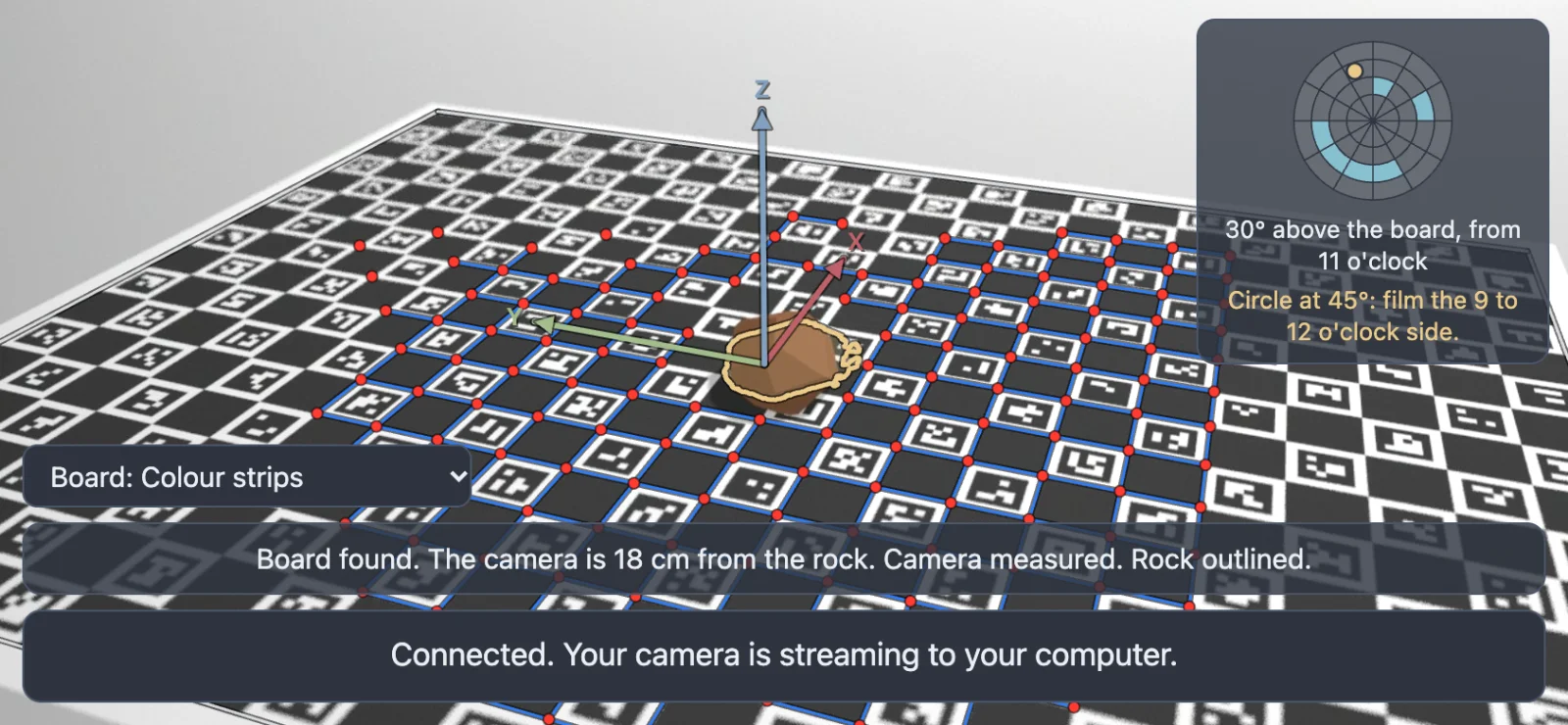 A phone held sideways over the board: red points on the corners of the printed squares it has found, joined by blue lines, red, green and blue arrows on the middle of the board, a yellow outline around the rock, a small round map with the camera's angle and a tip at the top right, and two lines of text at the bottom.